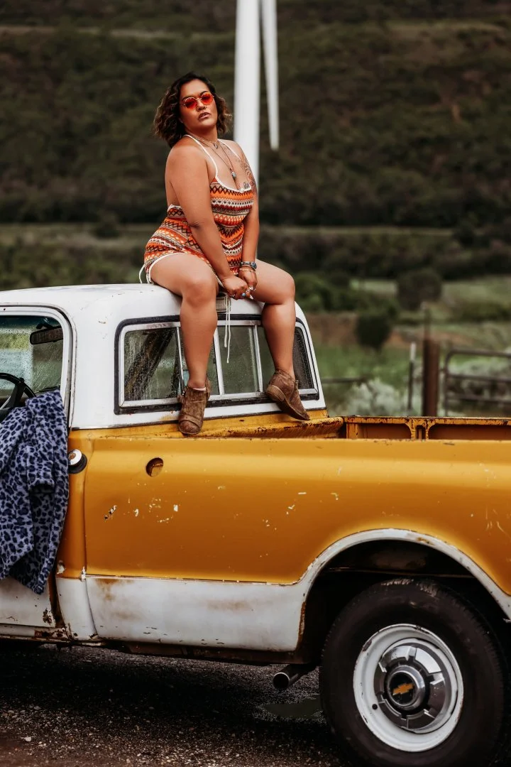 Woman with curly hair wearing sunglasses and a colorful striped dress sitting on the edge of a yellow and white pickup truck in a rural area.