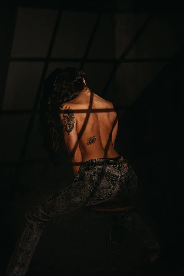 A woman with dark hair and tattoos on her back is sitting on a stool in a dark space. Light through a grid casts shadows on her back and shoulders.