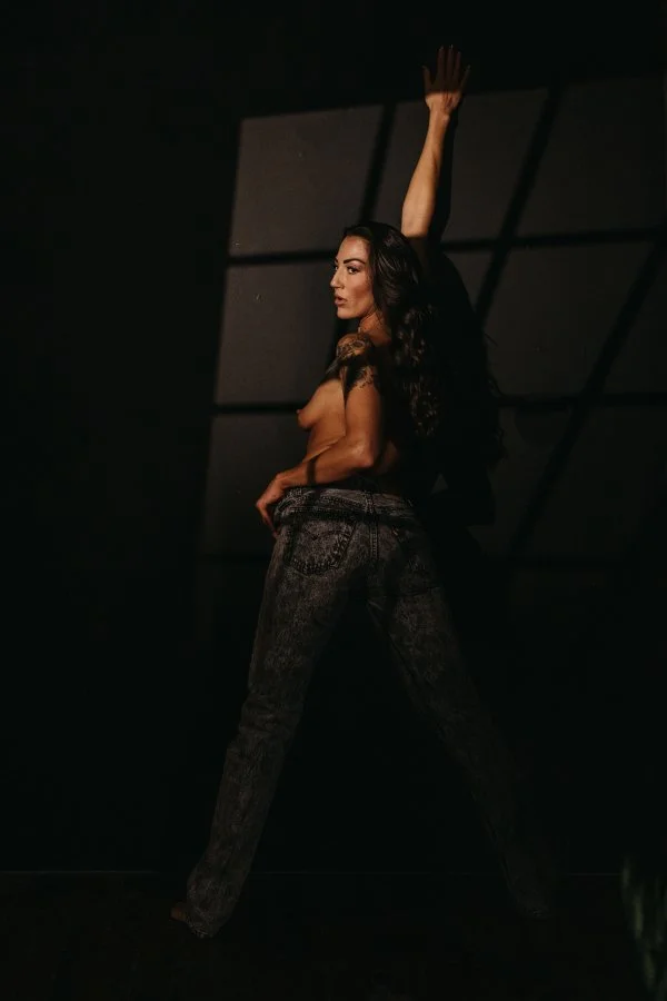 A woman with long dark curly hair standing in a dark room. She is topless, wearing high-waisted dark patterned jeans, with her right arm raised above her head and her left hand resting on her hip, looking to the side.