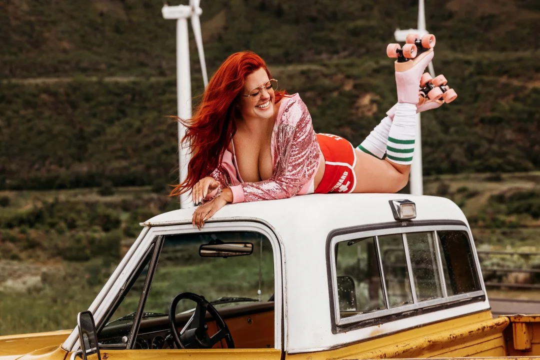 A woman with long red hair laying on top of a vehicle, smiling, holding skateboards, wearing a pink jacket, red shorts, and knee-high striped socks, outdoors with wind turbines in the background.