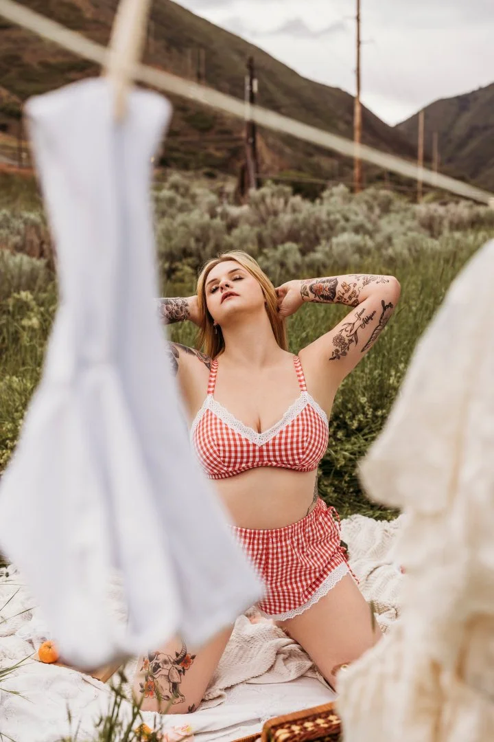 A young woman with tattoos rests on a blanket outdoors, wearing a red and white checkered crop top and shorts, with laundry hanging on a line in front of grassy hills and mountains.