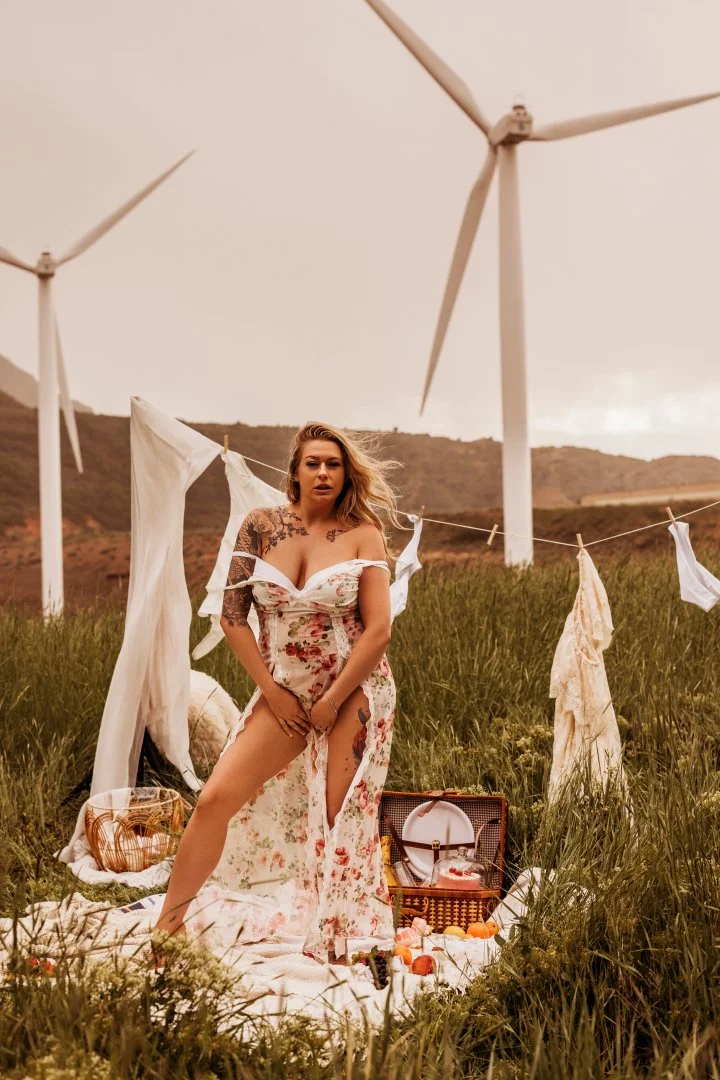 A woman in a floral dress standing in a grassy field with wind turbines in the background, surrounded by hanging white cloths and picnic items.