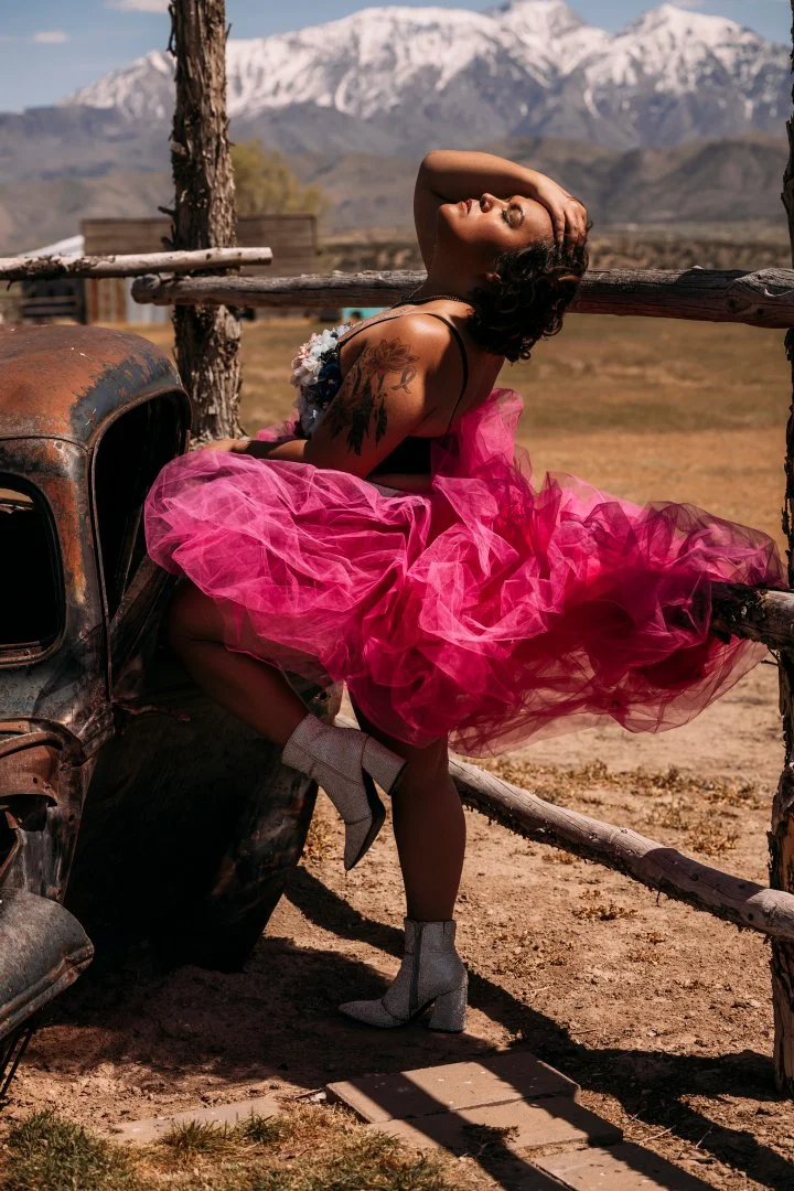 A woman in a pink tulle dress and high-heeled boots posing outdoors next to a rusted vintage car, with mountains in the background and a wooden fence nearby.