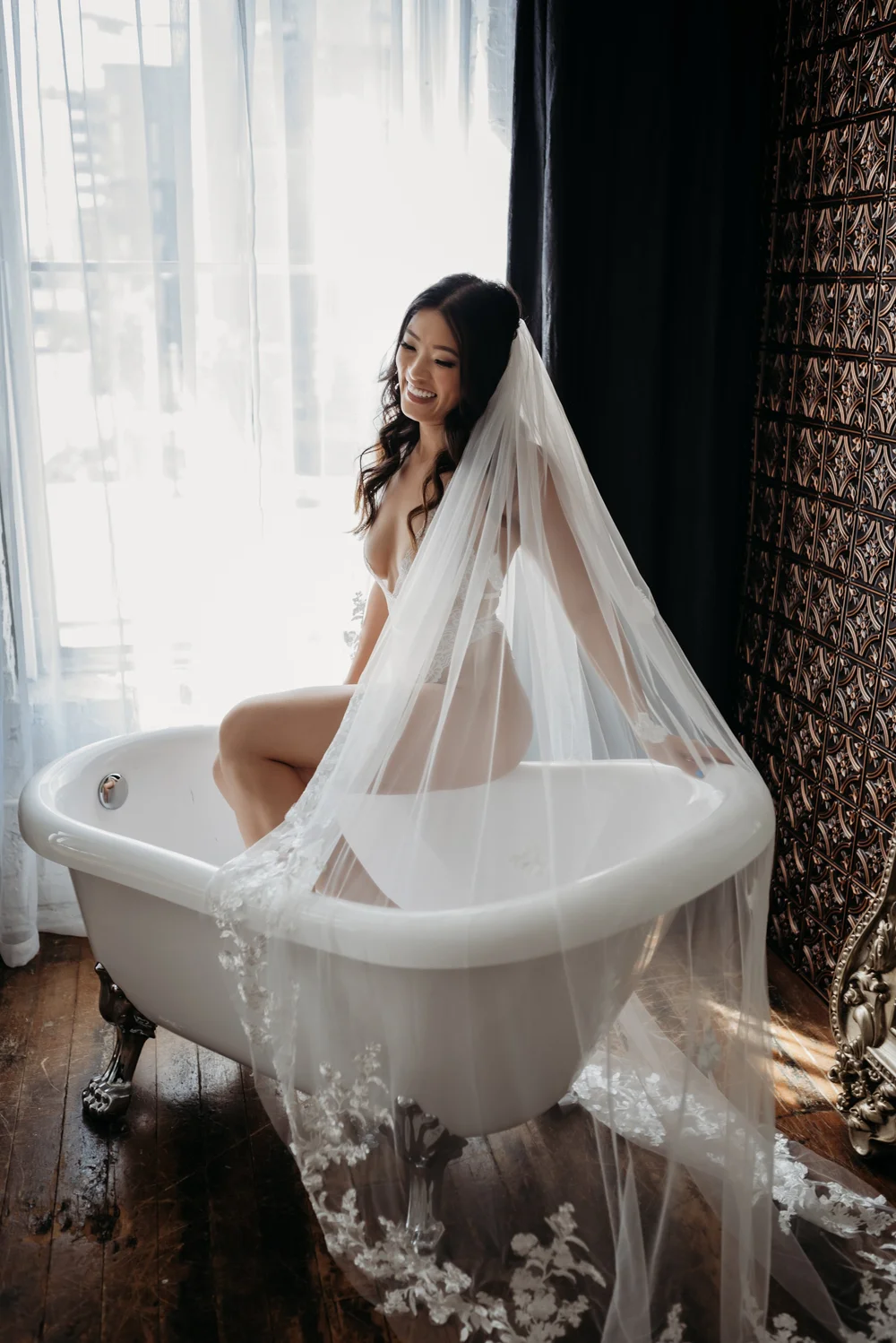 A smiling bride with a sheer veil in a white vintage bathtub near a window