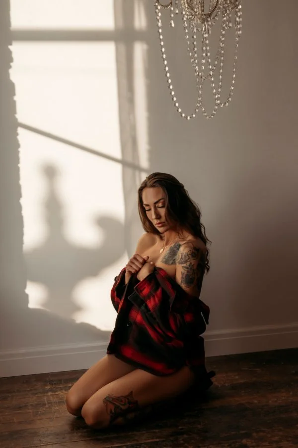 A woman kneeling on a wooden floor in a sunlit room, wearing an open red and black flannel shirt, with a tattoo on her right thigh and upper arm, and a tattoo on her chest. She has long dark hair and is looking down while holding her shirt. A chandel