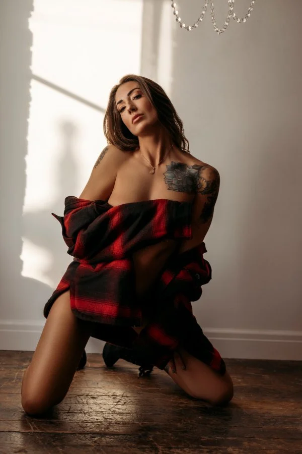 A woman with tattoos and shoulder-length hair, kneeling on the wooden floor in a softly lit room, wearing a red and black plaid shirt draped over her shoulders.