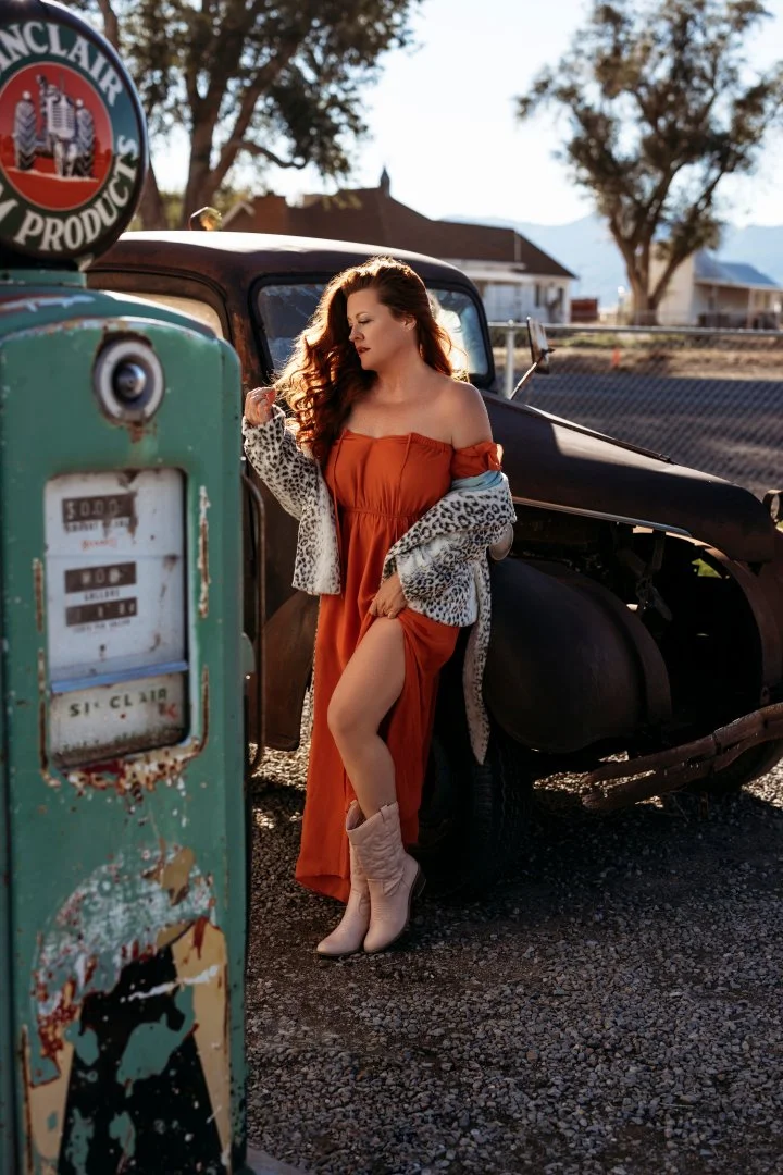 A woman with long, wavy hair wearing an orange dress, a leopard print jacket, and beige cowboy boots, leaning against an old black car. She is at a vintage gas station with an old, rusty gas pump in the foreground, and a sign reading 'McClair' in par