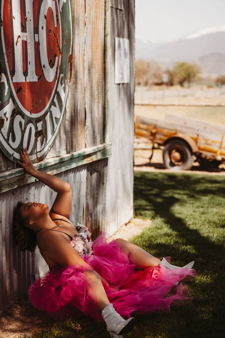 A woman wearing a pink dress and silver boots is sitting on the ground, leaning against a rustic wooden structure with a faded sign on it. She is smiling with her eyes closed and reaching up with one hand towards the sign.