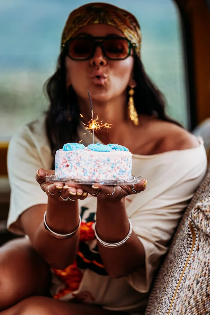 Woman wearing sunglasses, a headscarf, and jewelry holding a birthday cake with a sparkler, indoors or in a vehicle.
