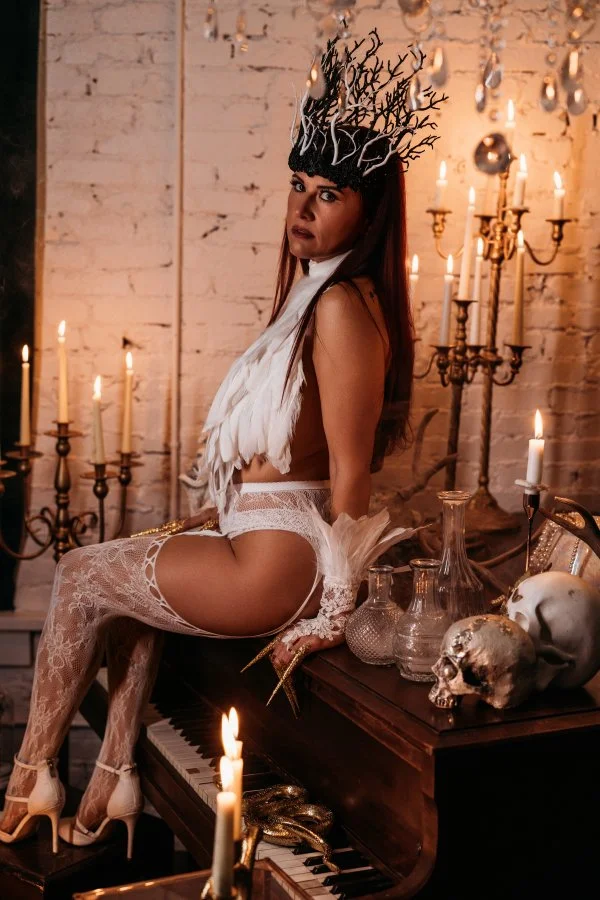 White feather fallen angel with antler headpiece seated at a candlelit piano, Utah Halloween boudoir