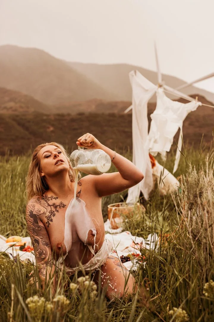 A woman with tattoos on her arms and shoulder, kneeling in a grassy field, pouring milk from a glass jar. She is partially covered by a white cloth. In the background, there is a clothesline with white laundry hanging, and mountains in the distance u