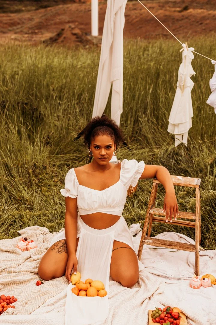 A woman in a white dress with cutouts kneels on a blanket in a grassy field, surrounded by fruits and desserts during sunset, with a wooden ladder, white drapes, and a clothesline in the background.