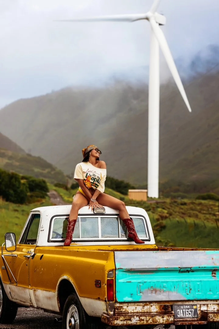 A woman sitting on top of a vintage yellow pickup truck, wearing cowboy boots, sunglasses, and a casual T-shirt, with a wind turbine and mountains in the background.