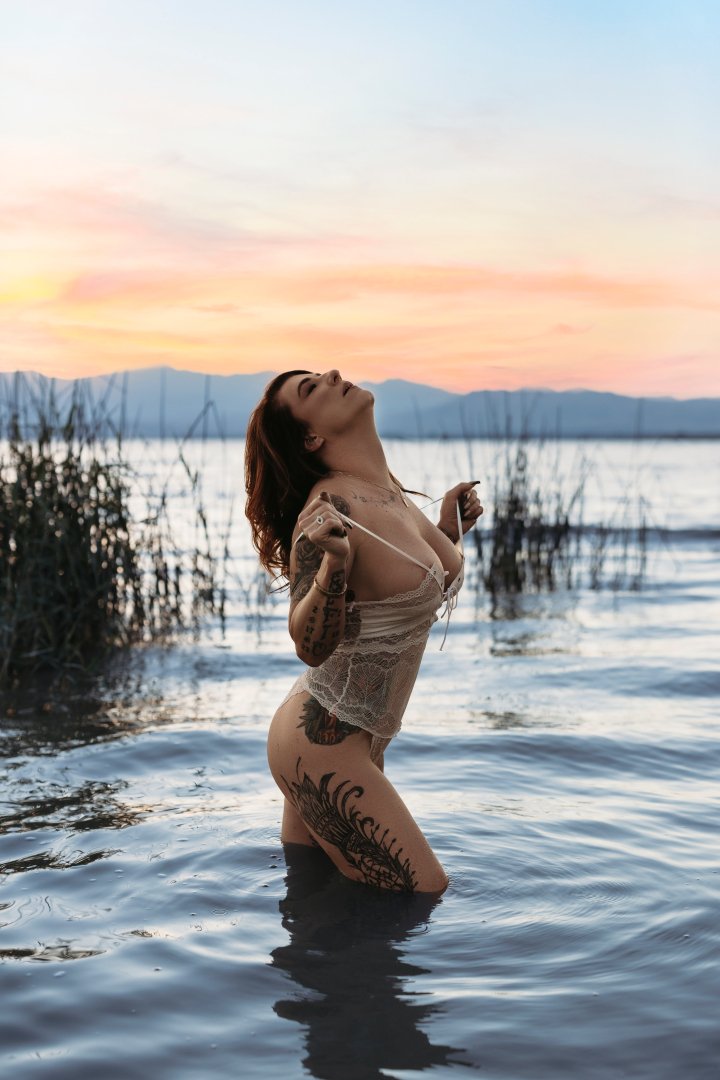 A woman with tattoos stands in a lake at sunset, wearing a lace top and holding a string, with mountains and colorful sky in the background.