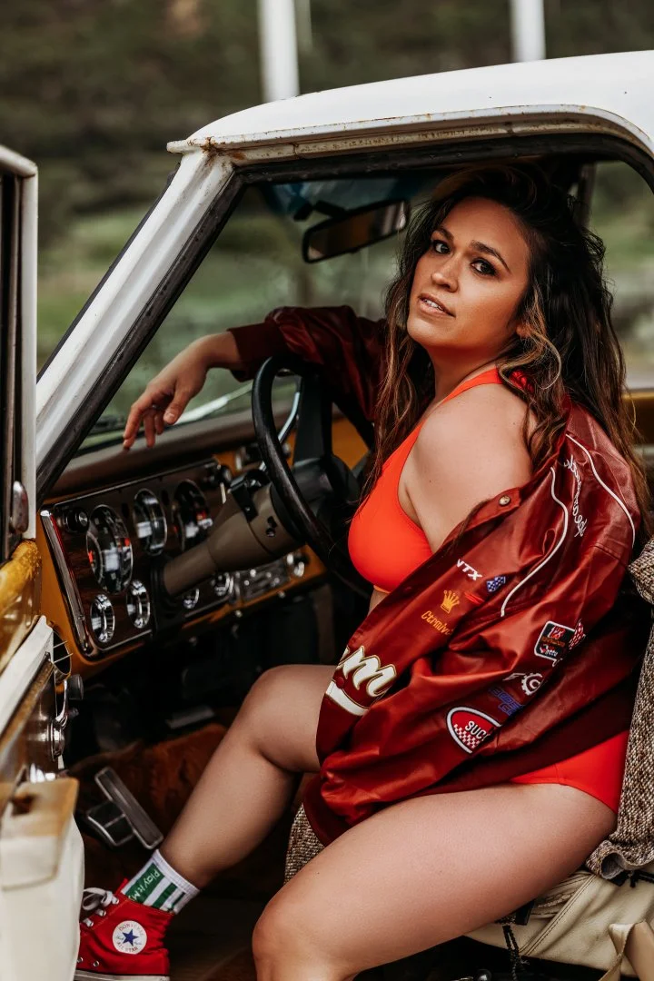 A woman wearing a red sports bra and a satin jacket with various patches, sitting in the driver's seat of a vintage car, looking at the camera.