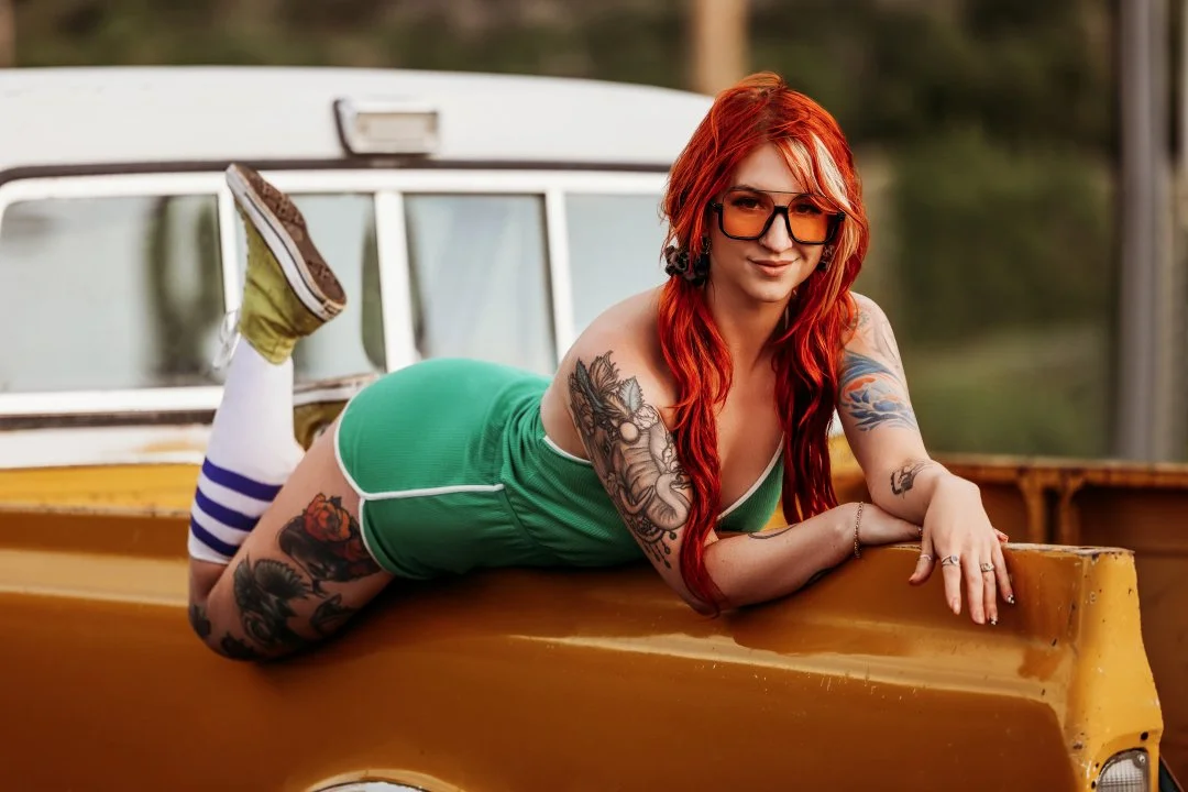 A young woman with red hair and tattoos, wearing sunglasses and a green tank top with striped socks, is lying on her stomach on the back of a yellow pickup truck.