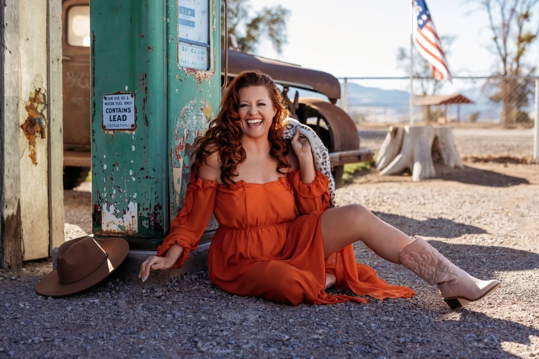 A woman with long curly red hair, wearing an orange off-shoulder dress and cowboy boots, sitting on the ground and laughing next to a vintage green gas pump in a rural setting.