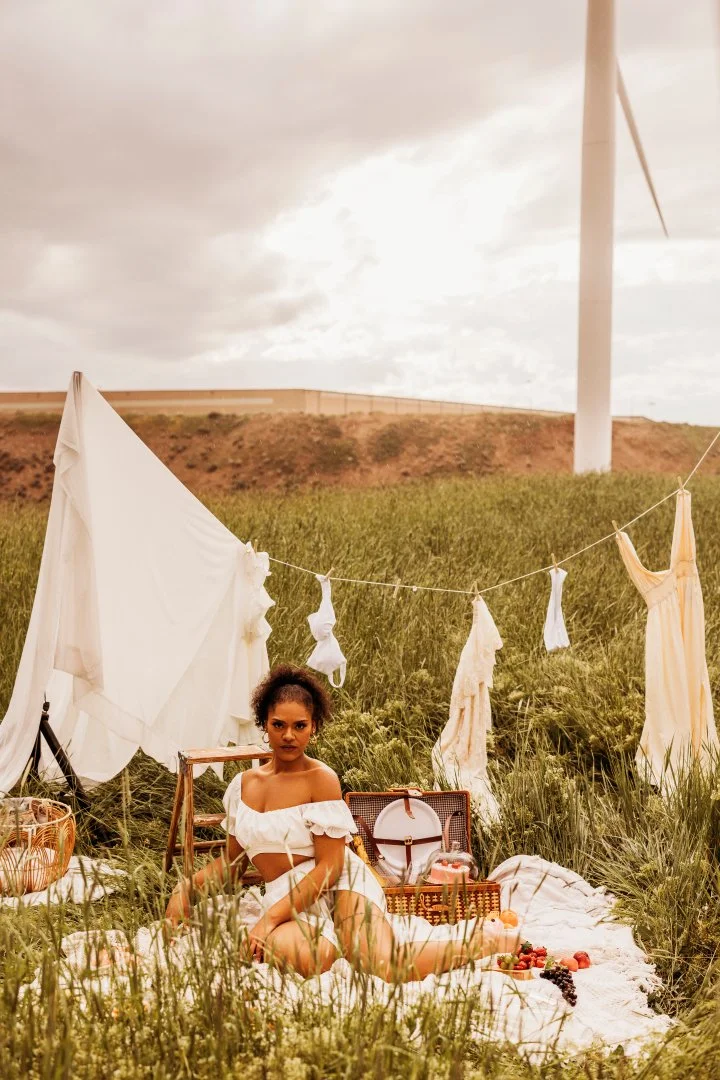 A woman in a white off-shoulder dress sitting on a blanket in a grassy field during daytime, with a wind turbine in the background, surrounded by hanging white clothes and a picnic setup with a wicker basket, a vintage suitcase, and food.