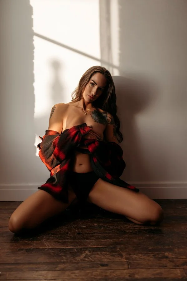 A woman with long dark hair, tattoo on shoulder, wears black underwear and an open red and black plaid shirt, sitting on a wooden floor with back against a wall, partially illuminated by sunlight through a window.