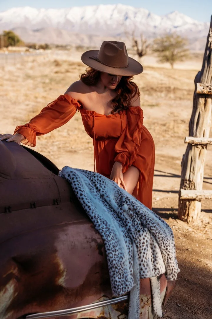 A woman in a brown hat and orange dress leaning on a vintage car covered with a leopard-print blanket in a desert landscape.