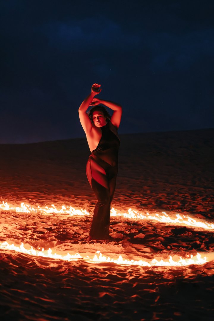 A woman in a black outfit performing a fire dance or fire ritual in a desert at night, surrounded by circular fire patterns.
