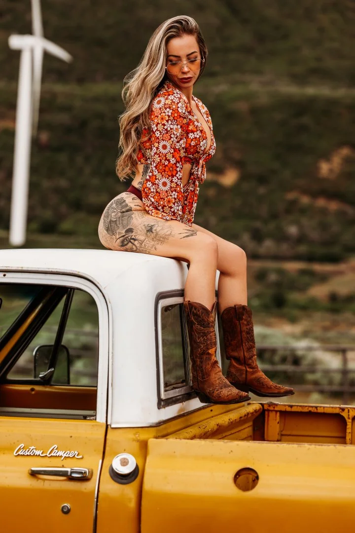 A woman with long wavy blonde hair wearing sunglasses, a red floral crop top, and cowboy boots sitting on the back of a yellow vintage truck with a white cab, outdoors in a hilly landscape.