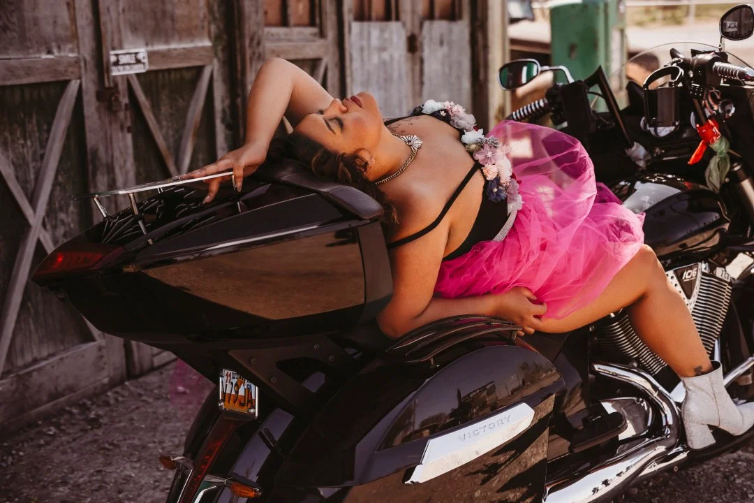 Woman lying on custom black motorcycle, wearing a pink tulle skirt, black top, flower necklace, and white boots, with her eyes closed and relaxed.
