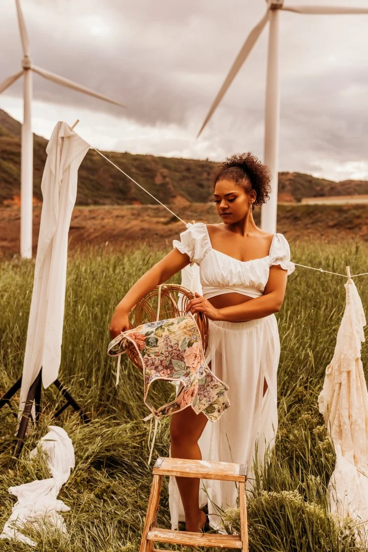 A woman in a white dress hanging laundry in a field of tall grass, with wind turbines and hills in the background under cloudy skies.