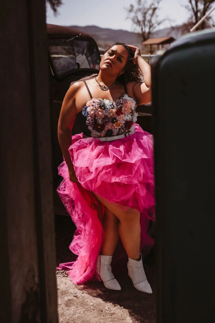 A woman in a floral top and a pink tulle dress with white boots standing outdoors near cars.