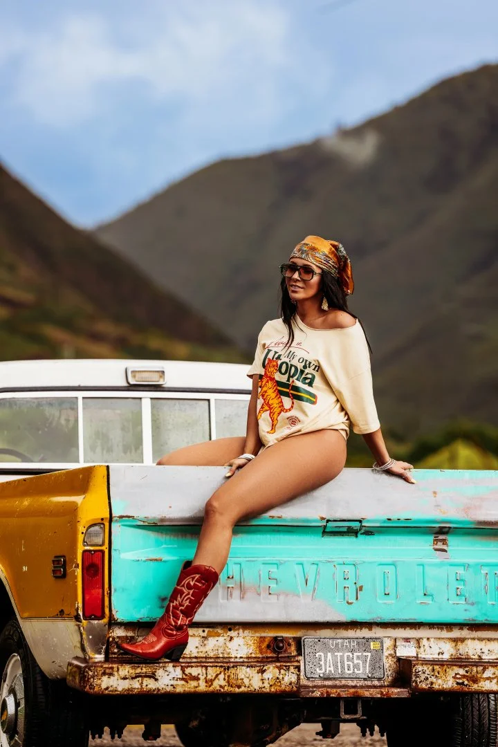 A young woman sitting on the tailgate of a vintage Chevrolet pickup truck in a mountainous outdoor setting. She is wearing a bandana, sunglasses, a graphic T-shirt, and cowboy boots.