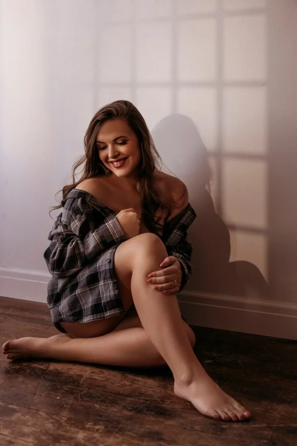 A woman sitting on the floor with her back against the wall, wearing a plaid shirt off her shoulders, smiling and holding one knee, with sunlight casting a shadow on the wall.