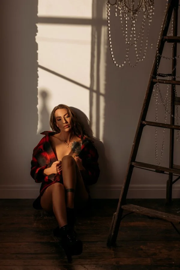 A woman with long hair sitting on the floor against a wall, wearing a red and black checkered shirt, black stockings, and black shoes. She is holding her knee with one hand. Behind her, sunlight casts shadows of window frames and a chandelier on the 