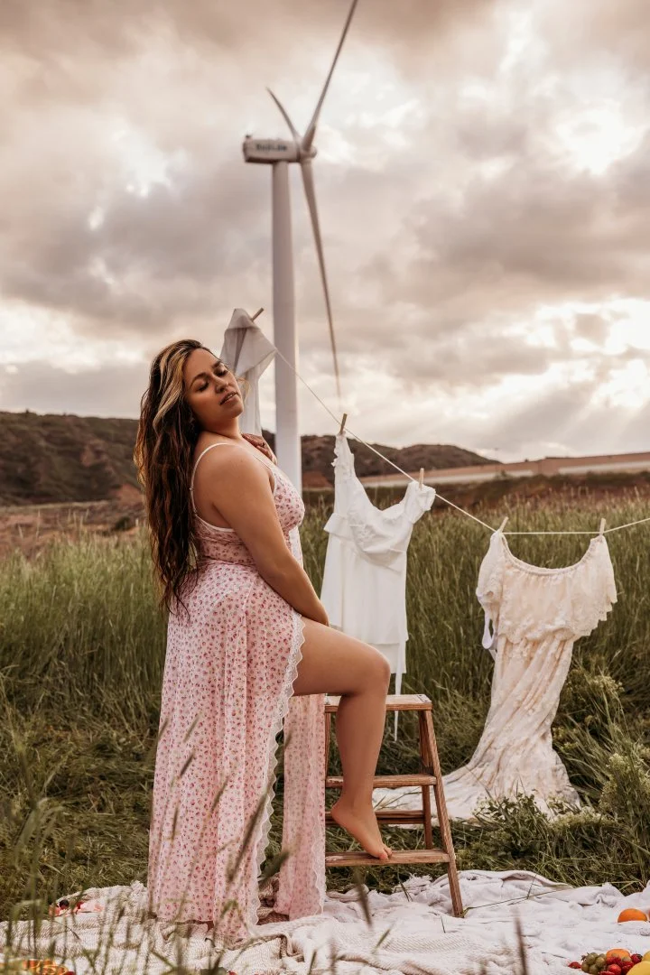 A woman in a pink floral dress kneeling on a step stool in a field with a wind turbine in the background. Clothes are hanging on a clothesline behind her, and she appears to be posing with her eyes closed during sunset or sunrise.