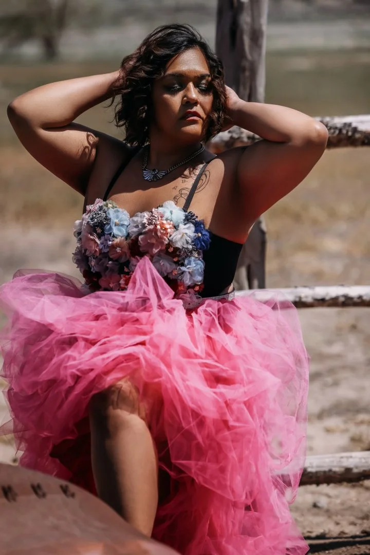 A woman in a pink tulle skirt with a floral top, sitting outdoors with her hands behind her head, looking down.