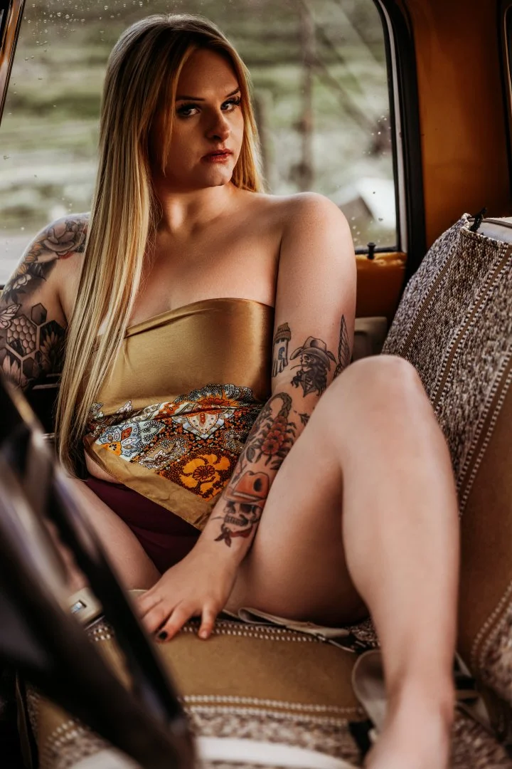 A young woman with long blonde hair and tattoos sitting inside a vehicle, wearing a strapless dress, looking at the camera with a serious expression.
