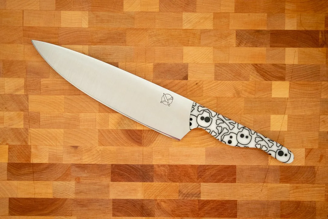 Blue Collar Blades 8" Chefs Knife with stainless steel blade and white resin with black skulls in the knife handle resting on a cutting board.