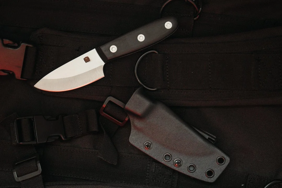 S35V stainless steel fixed-blade knife with black G10 handle and stainless steel bolts. Small stamped logo on the blade, a black kydex sheath, and tactical backpack in the background. 