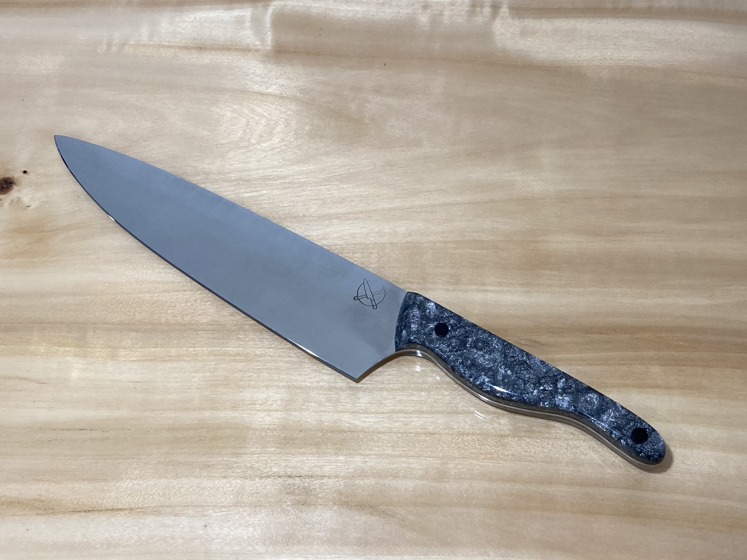 A kitchen chef's knife with a stainless steel blade and a black and gray speckled handle resting on a light wooden surface.