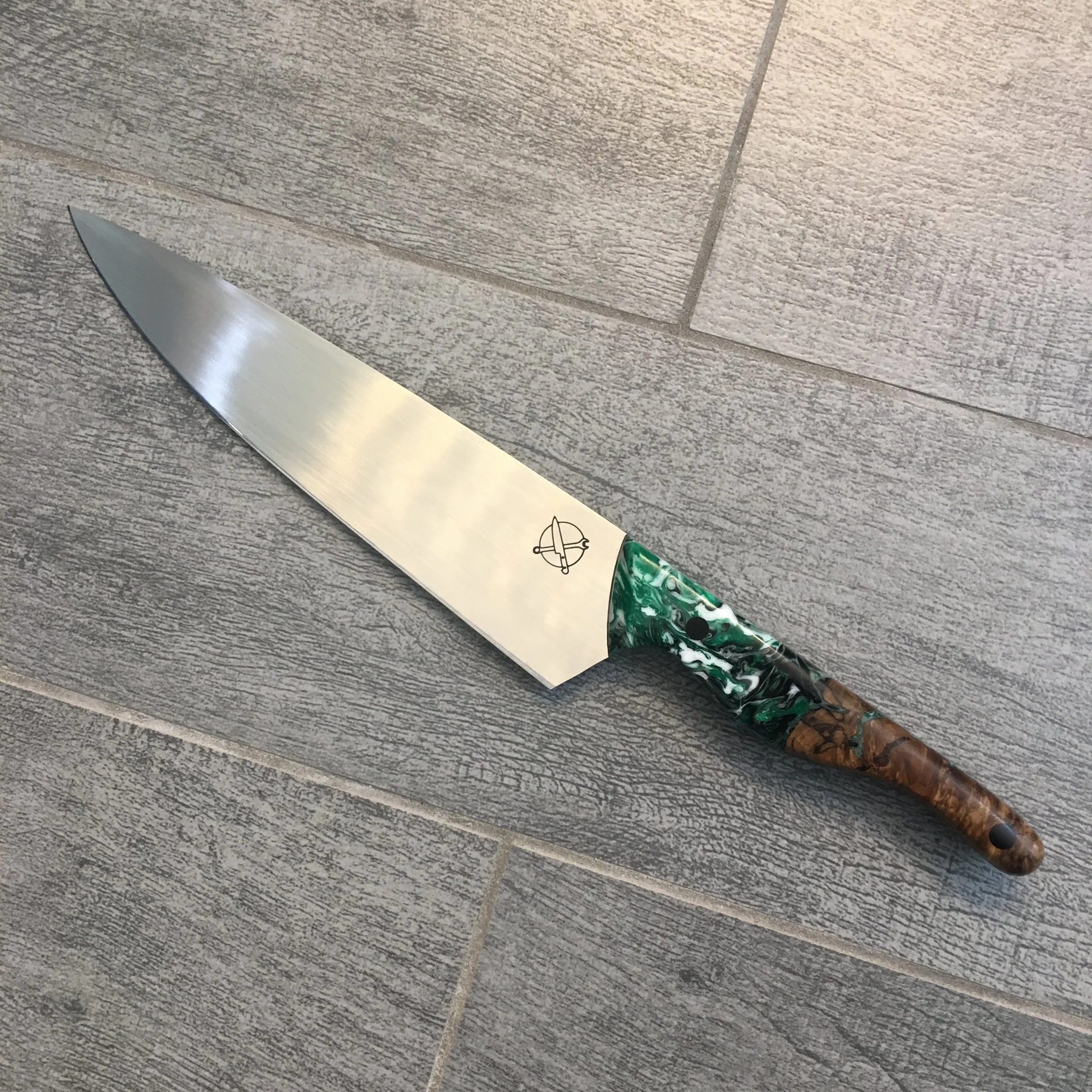 A kitchen knife with a wide stainless steel blade, decorated handle with green and brown marbled patterns, placed on a gray tiled floor.