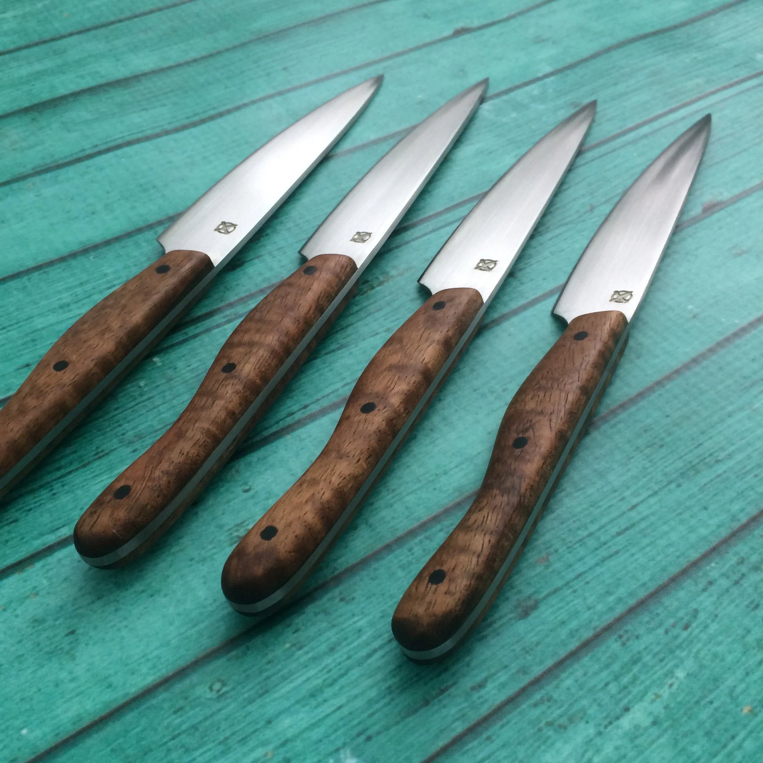 Four stainless steel steak knives with Hawaiian KOA wooden handles and carbon fiber pins arranged on a turquoise wooden surface.