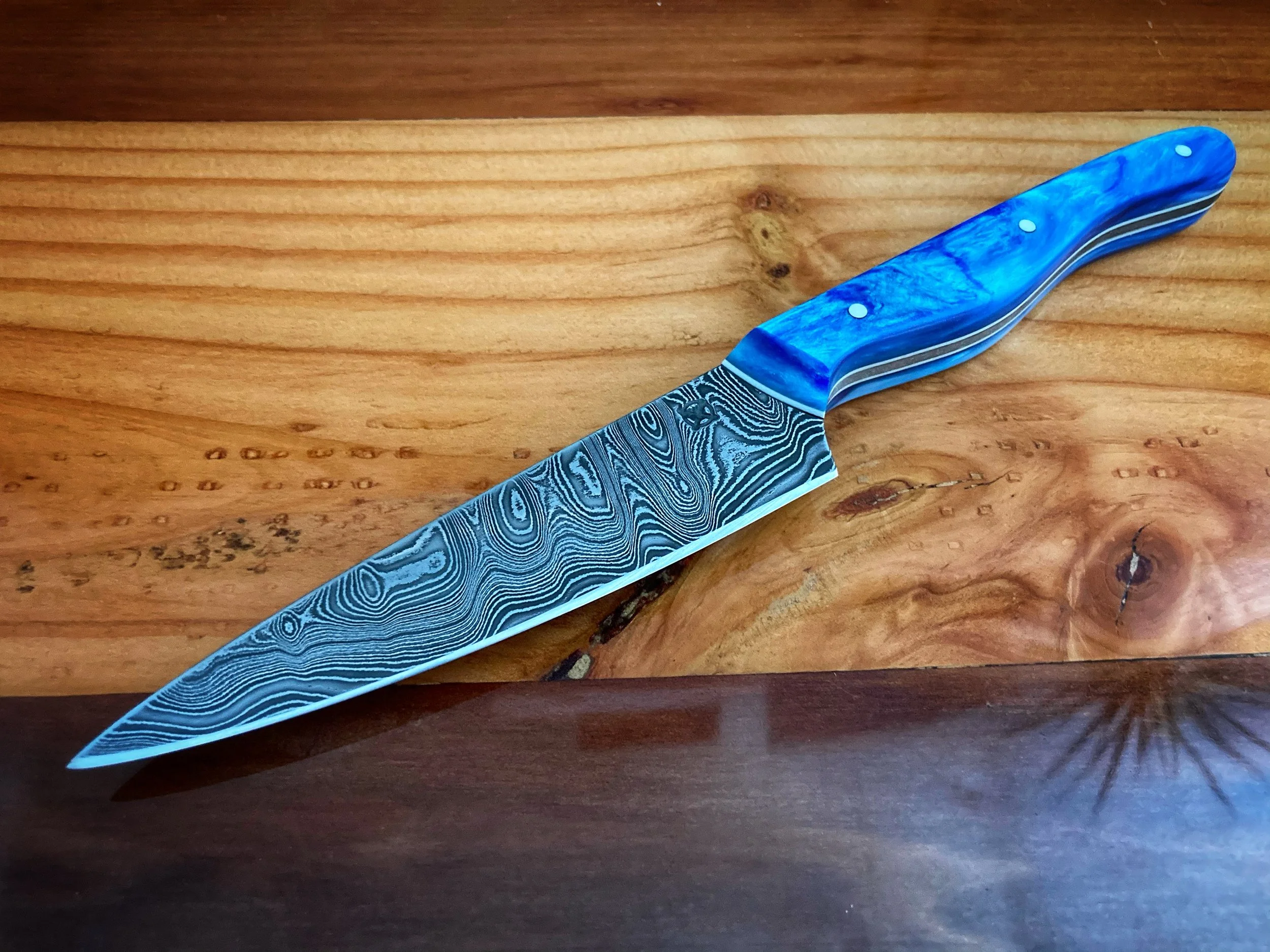 A Damascus steel kitchen knife with a blue handle resting on wooden surface.