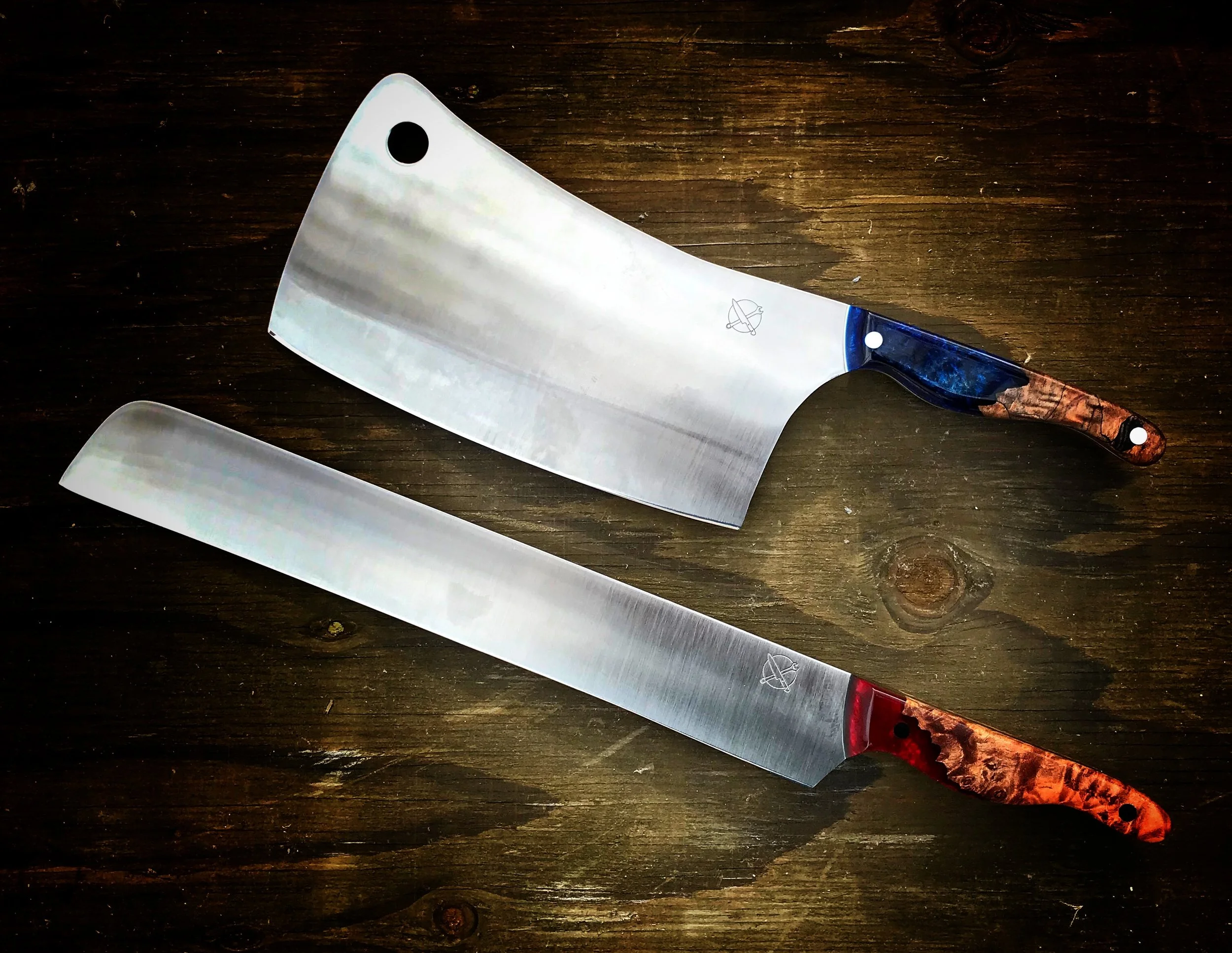 Two chef's knives with wooden handles on a dark wooden surface. The knives have broad blades with one curved and the other straight, used for slicing and chopping.