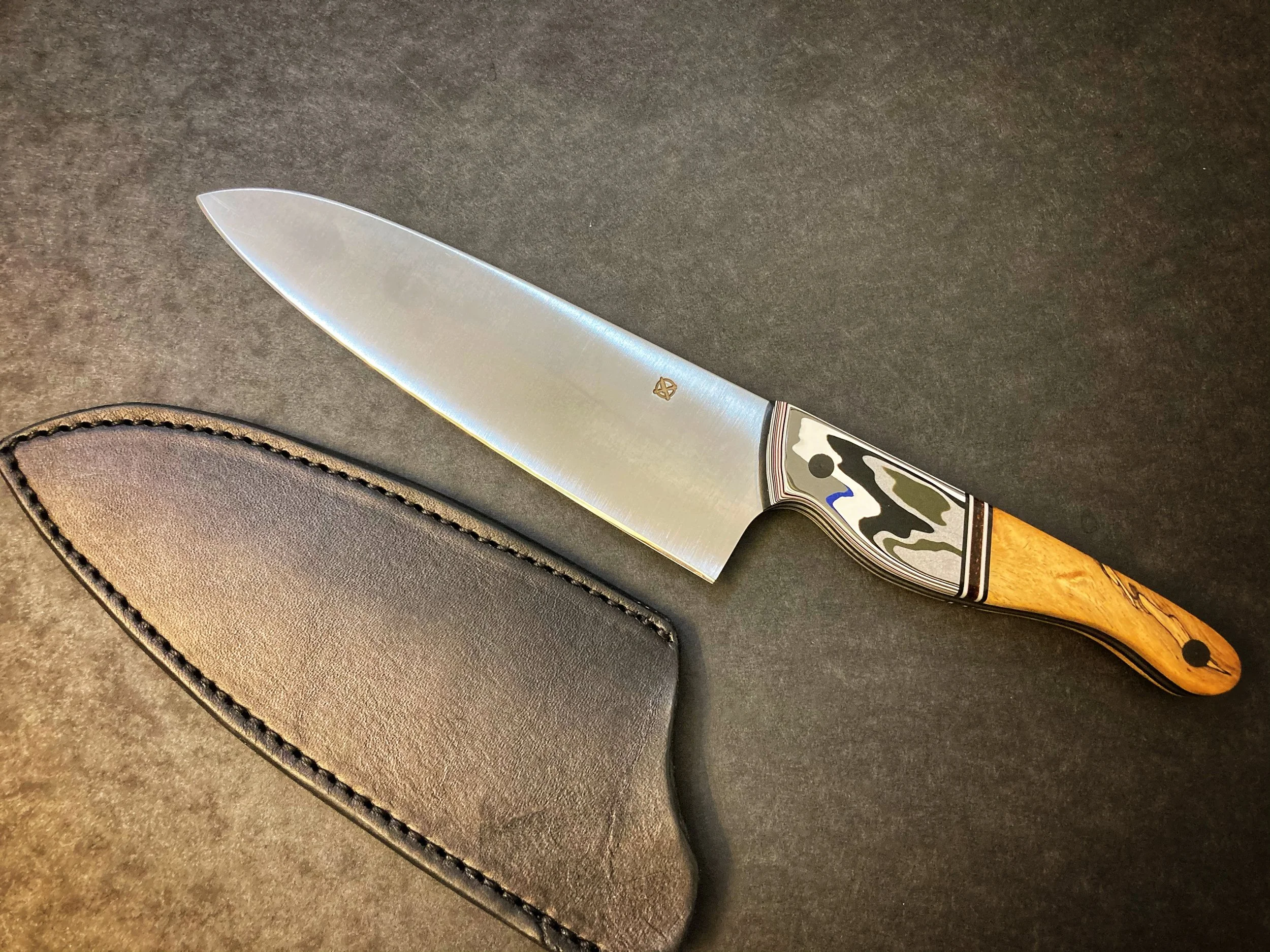 A hunting or kitchen knife with a decorative handle and a leather sheath on a dark surface.