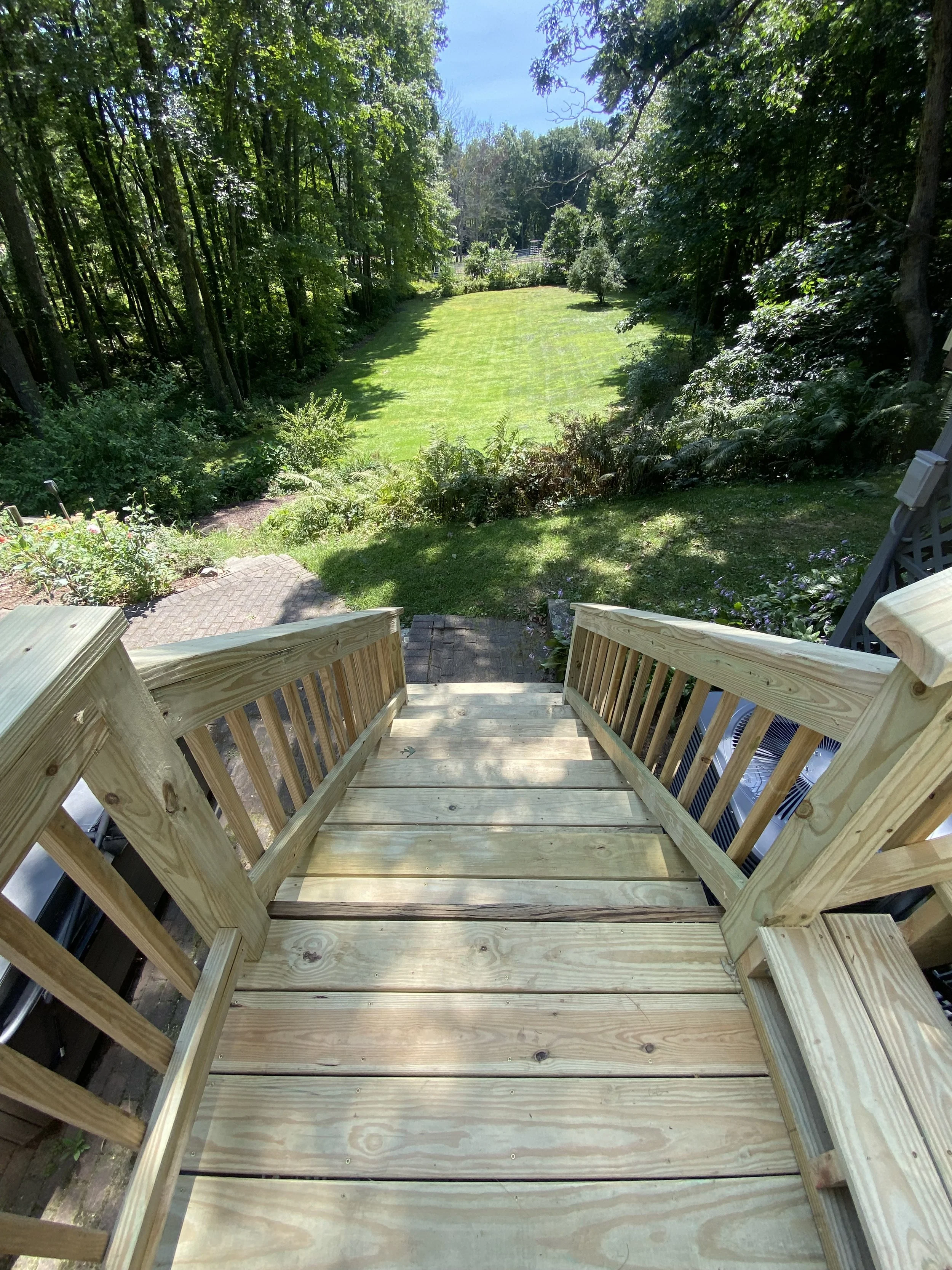 View from wooden stairs overlooking a backyard with a well-kept grassy lawn, trees, and lush greenery under a clear blue sky.