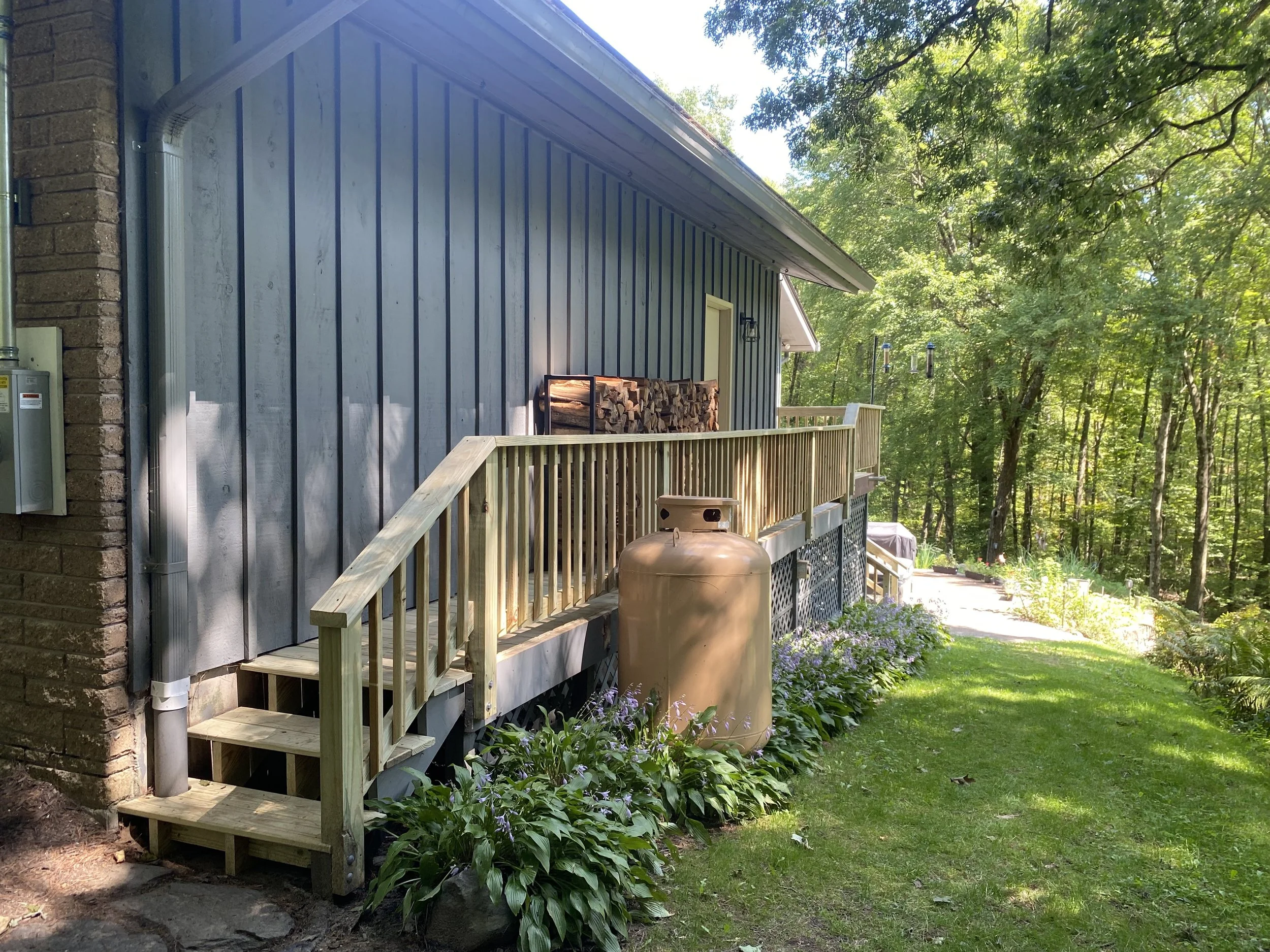 Side view of a house with a blue exterior, a wooden deck with stairs, a wood storage rack, and a propane tank, surrounded by green grass, plants, trees, and hanging wind chimes.
