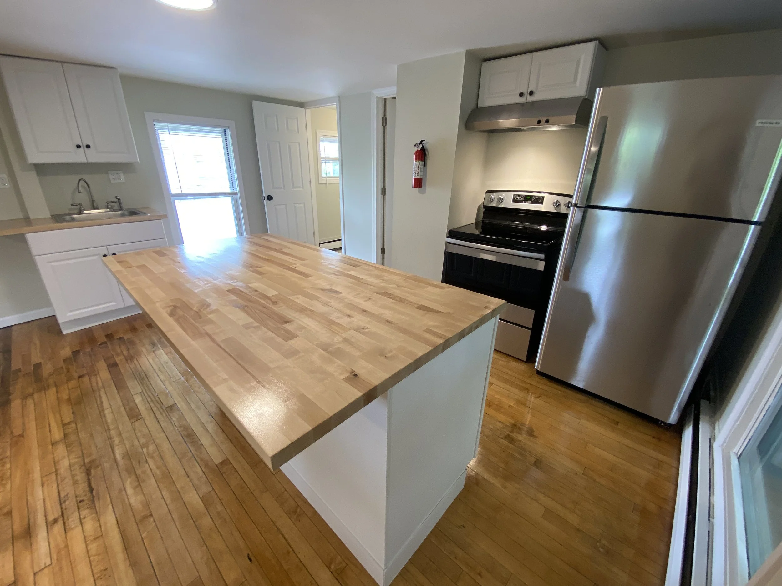 Kitchen with wooden countertop island, stainless steel refrigerator, black stove with stainless vent hood, white cabinets, sink under window, hardwood floors, door and window next to sink.