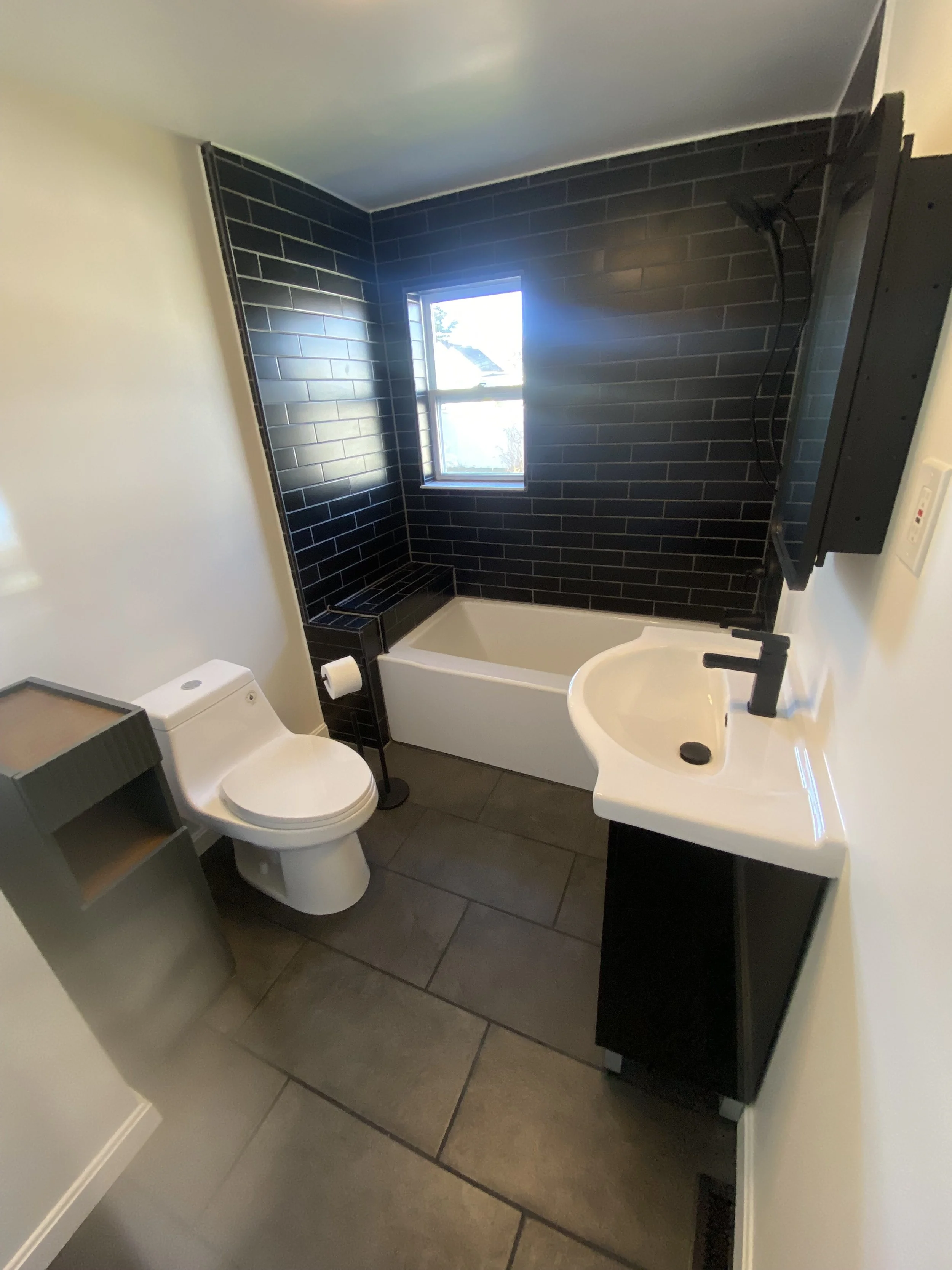 Small modern bathroom with black tile wall, white bathtub, white toilet, black and white sink, window, gray floor tiles.