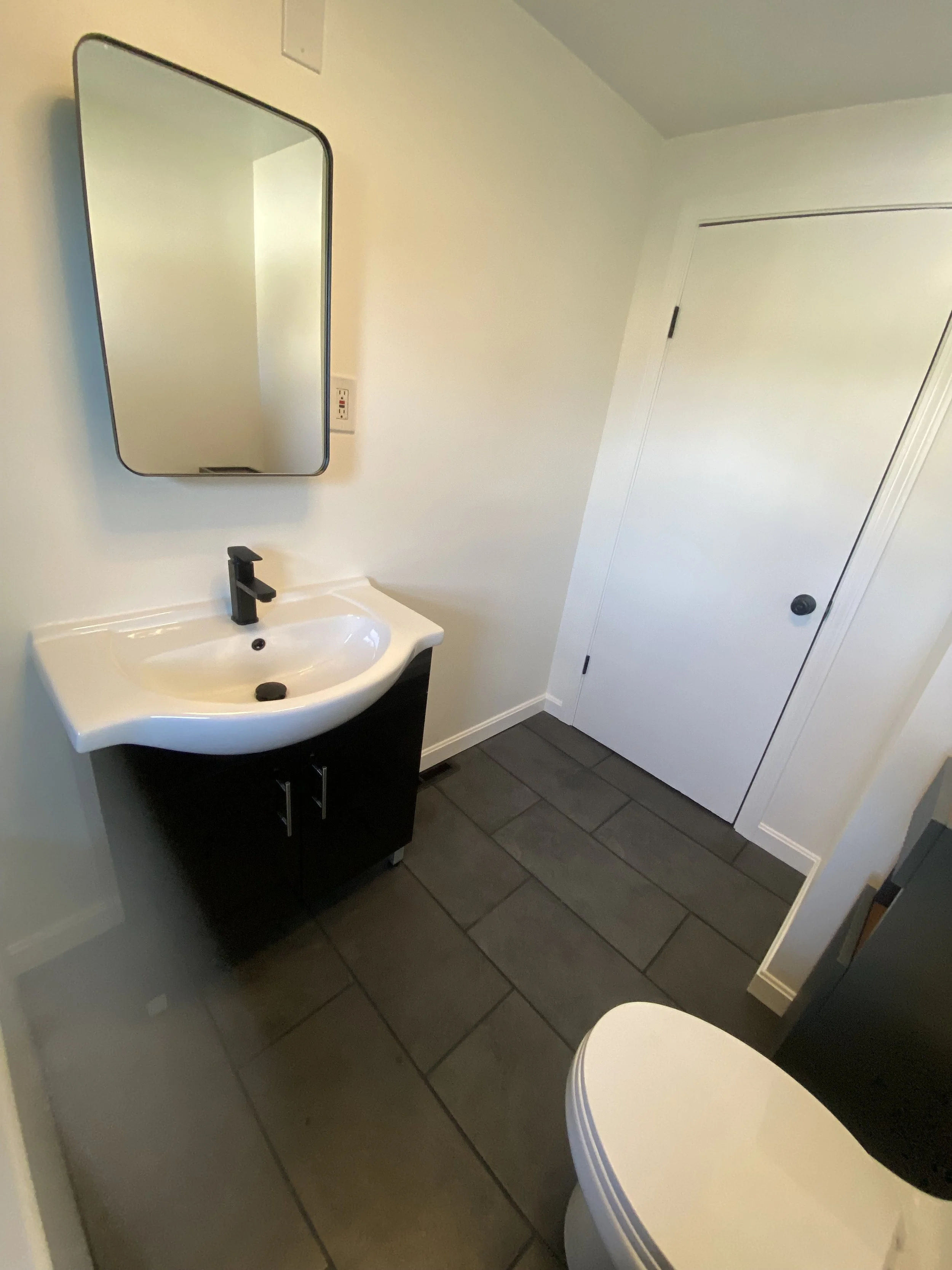 Small bathroom with a white sink and black vanity, a large mirror above the sink, a closed white door, and a portion of a toilet in the foreground. The flooring is dark tile.