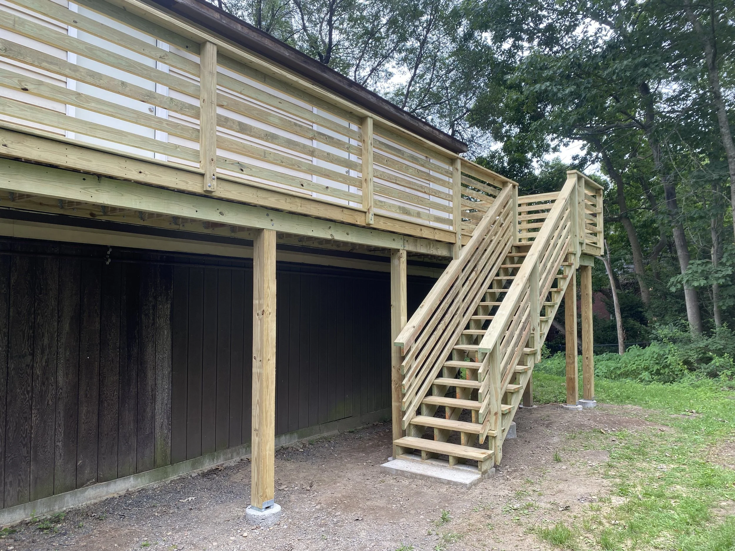 Newly built wooden outdoor staircase leading to a deck with a railing, set in a backyard with trees.