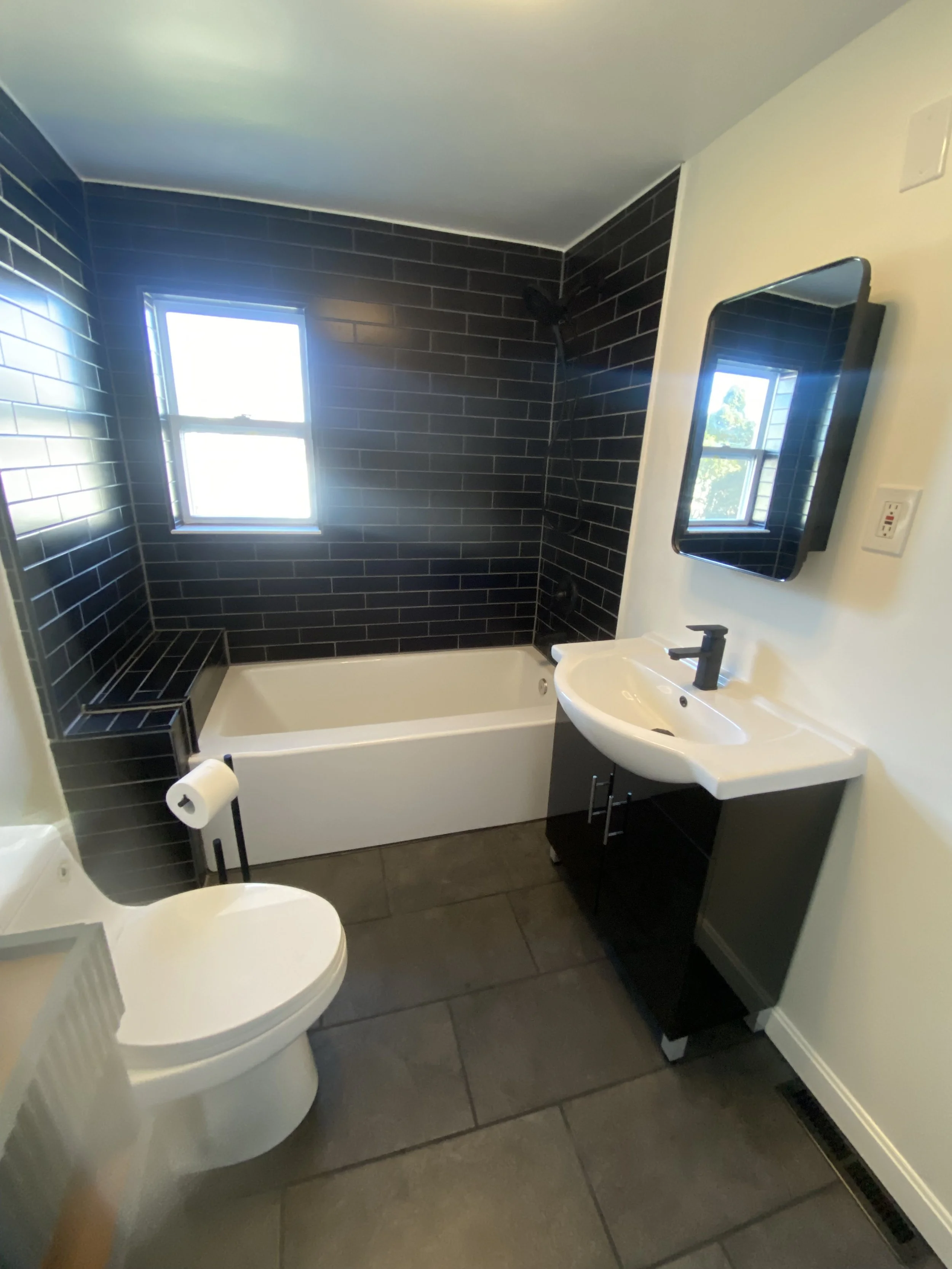 Small modern bathroom with dark gray tiled floor and black tiled walls around the bathtub, white bathtub, white toilet with a toilet paper holder, small white sink with gray cabinet, black faucet, and black-framed mirror above the sink. There is a wi